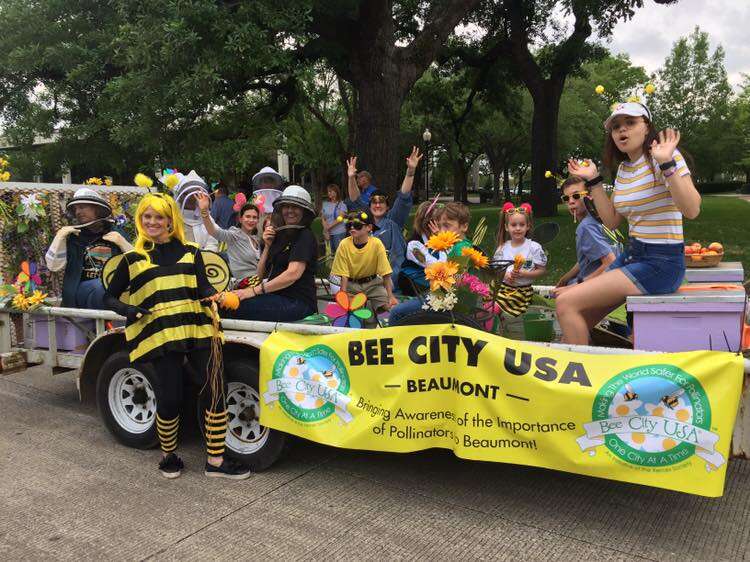A smiling group of kids and adults sit on a parade float with a yellow Bee City banner on the side, a woman in a bee costume stands in the foreground.