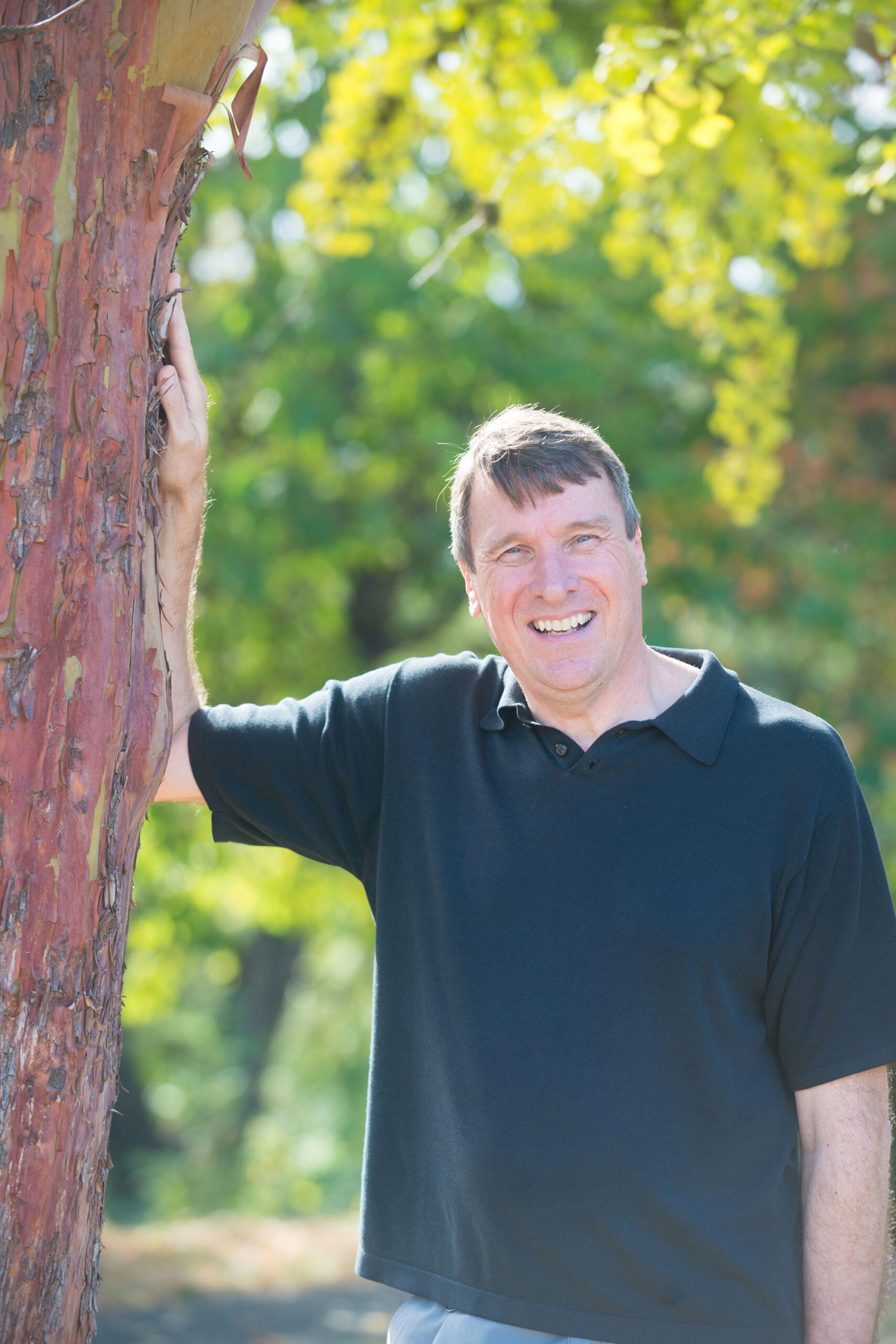 Man in black polo shirt smiling, next to tree