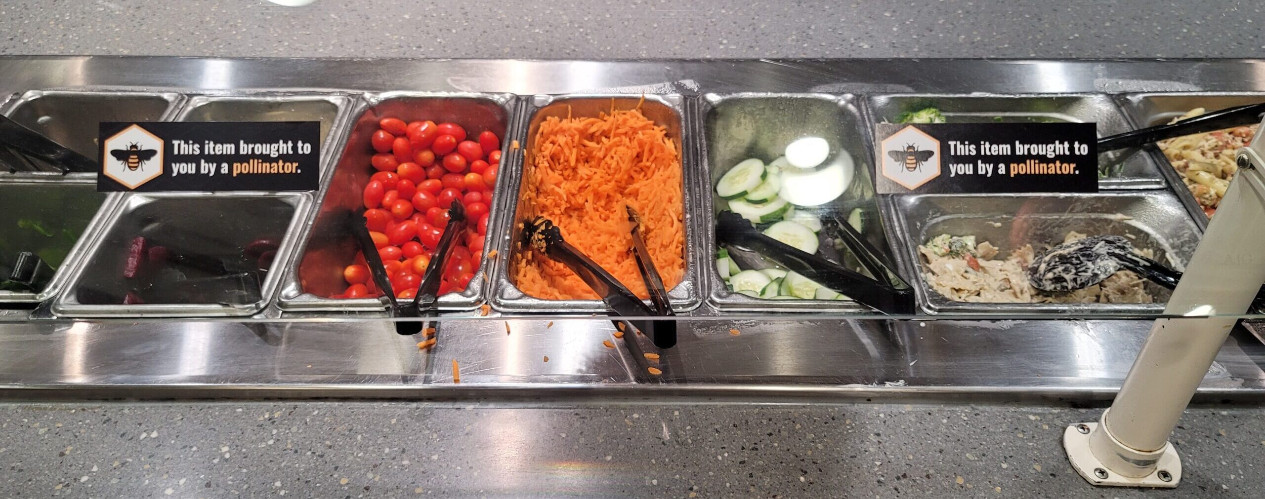 Vegetables in metal cafeteria trays with 2 stickers on glass guard saying "This item brought to you by a pollinator