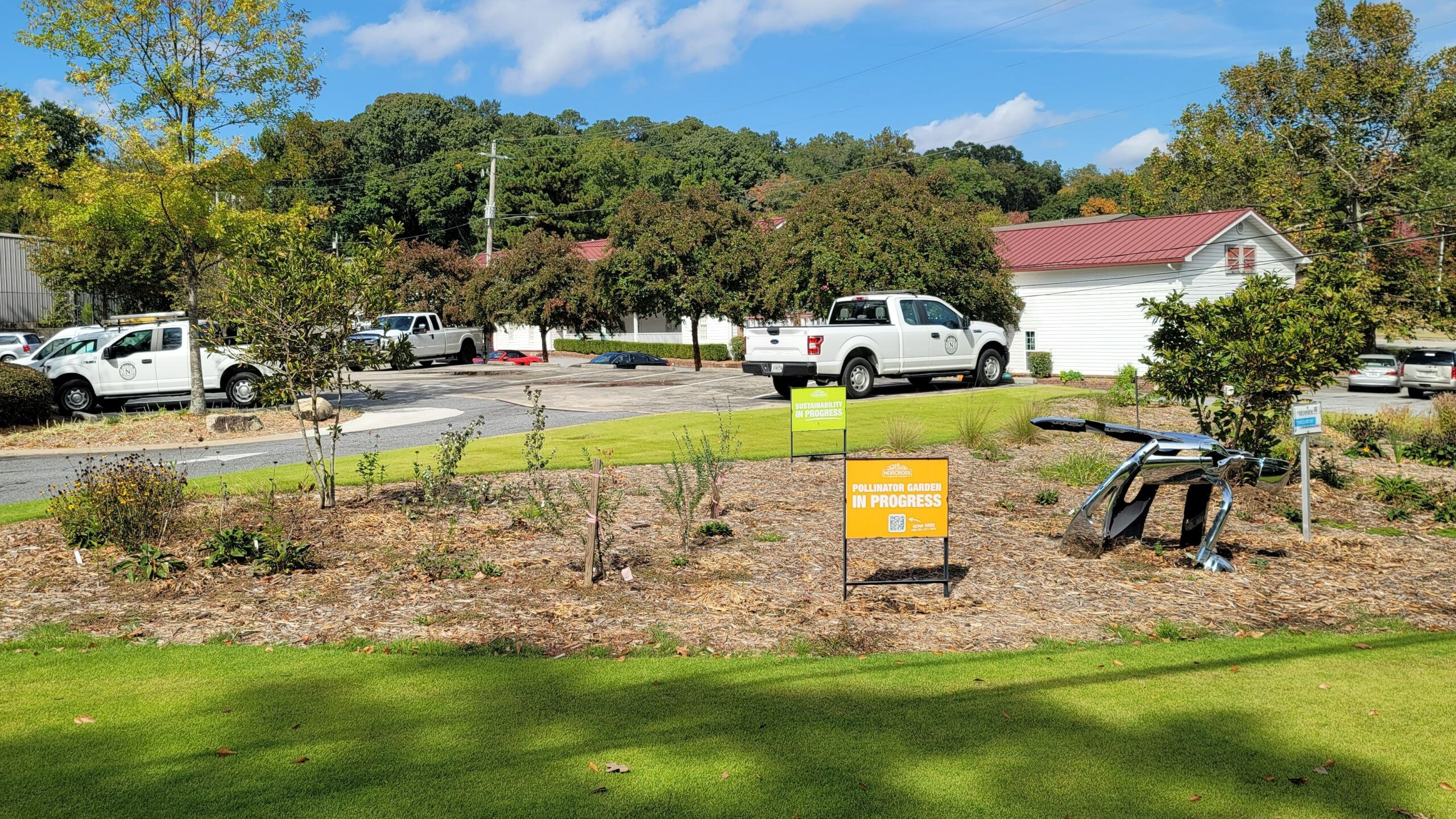 A mulched, planted area in a green lawn, next to a parking lot with a blue sky overhead.