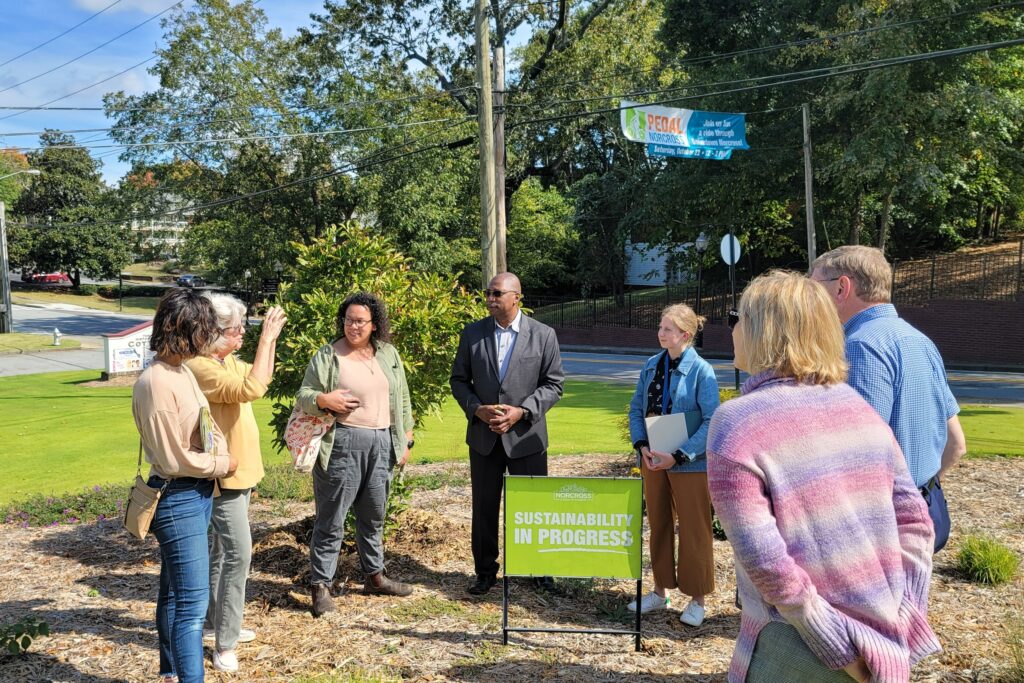 A circle of people listen to a speaker on a sunny day. A green yard sign in the middle of the group reads "Sustainability in Progress".