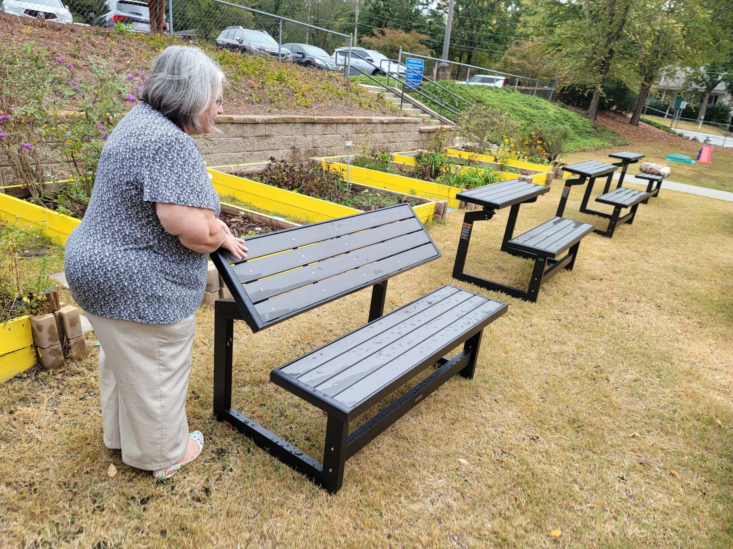 A woman in gray and tan clothes adjusts a gray bench that converts to a outdoor desk, with yellow raised garden beds in the background.