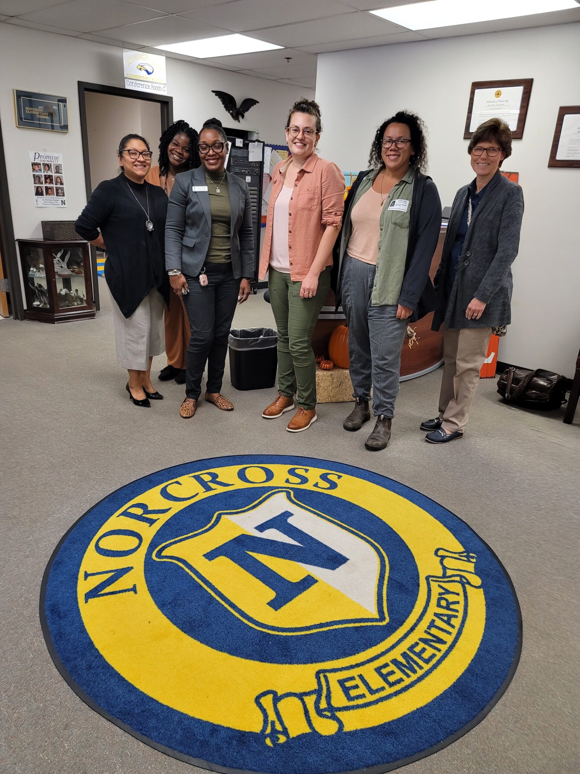 Six people stand in an office setting in front of a yellow and blue emblem on the carpet in front of them, which reads "Norcross Elementary".
