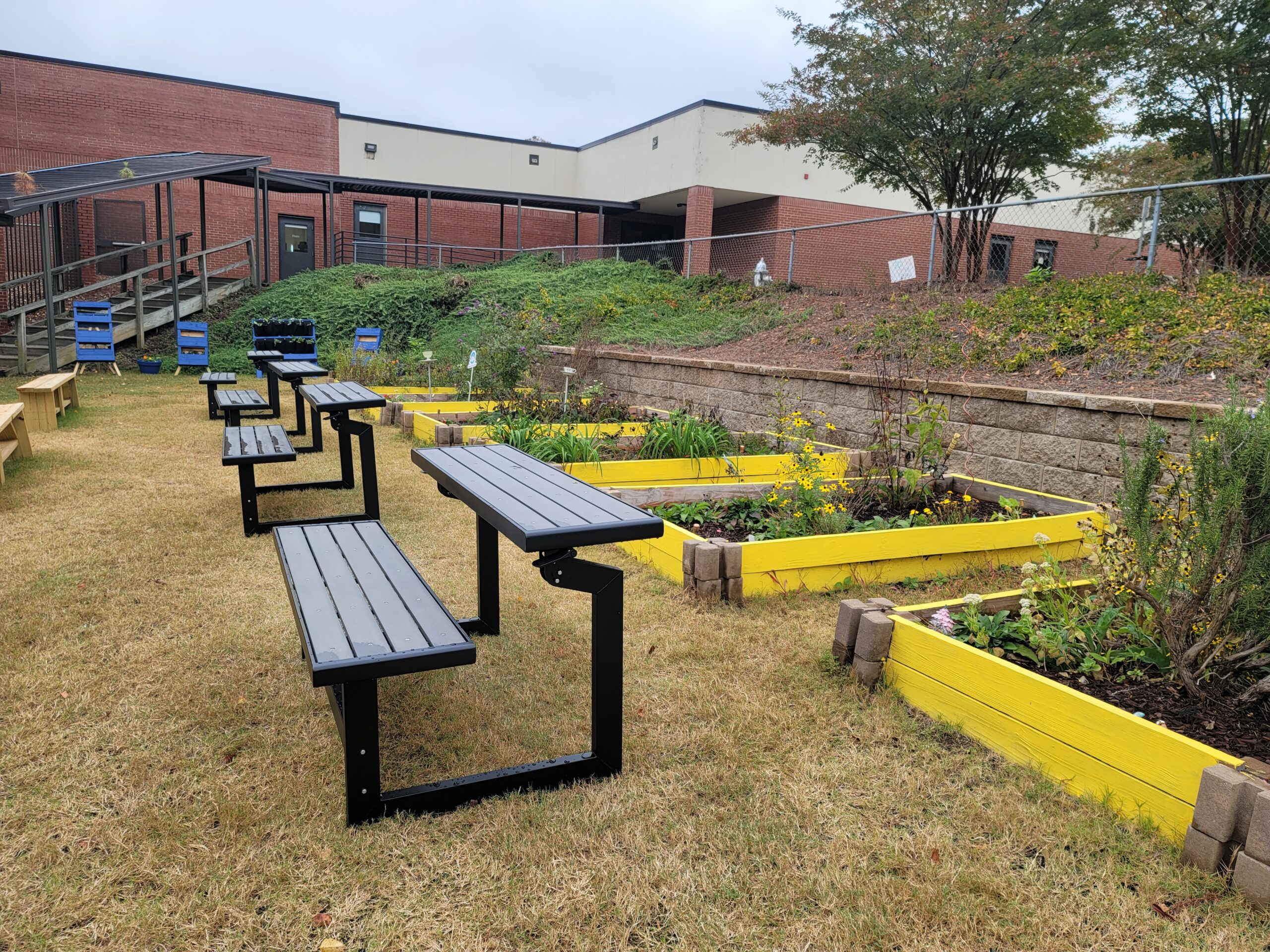 A row of gray outdoor desks face a row of yellow raised garden beds.