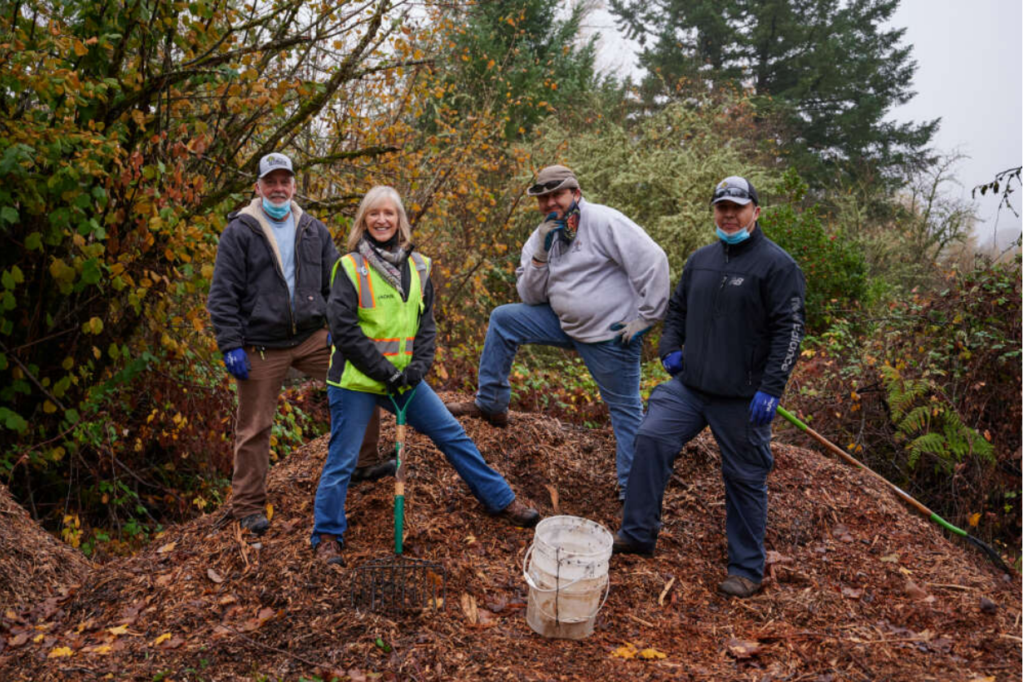 Four smiling adults in warm work clothes stand on a brown mulch pile with green trees and shrubs in the background.