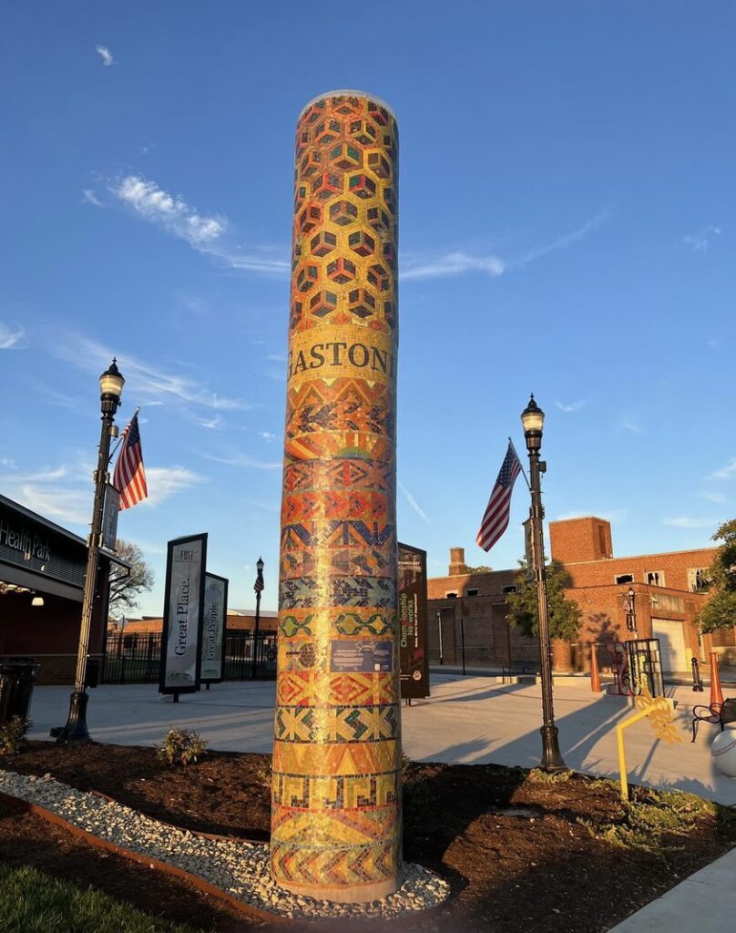The mosaic column shining in the sun, with blue sky in the background. In the background there is an open courtyard with lampposts and American flags, and a brick building in the far background.