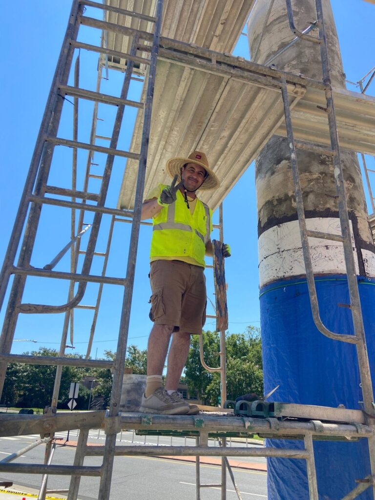 A person smiling and giving the peace sign stands on scaffolding in a high-viz vest, straw hat and shorts, with blue sky and a road in the background.
