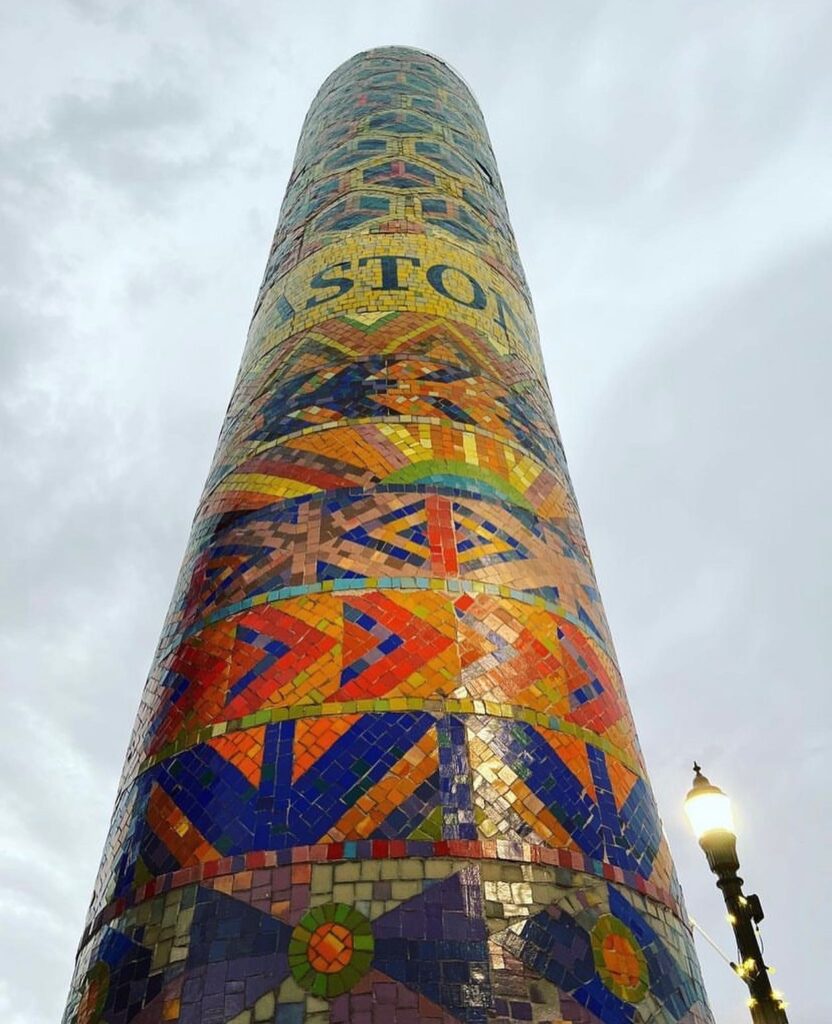 Looking up at a colorful mosaic column with a light gray sky in the background.