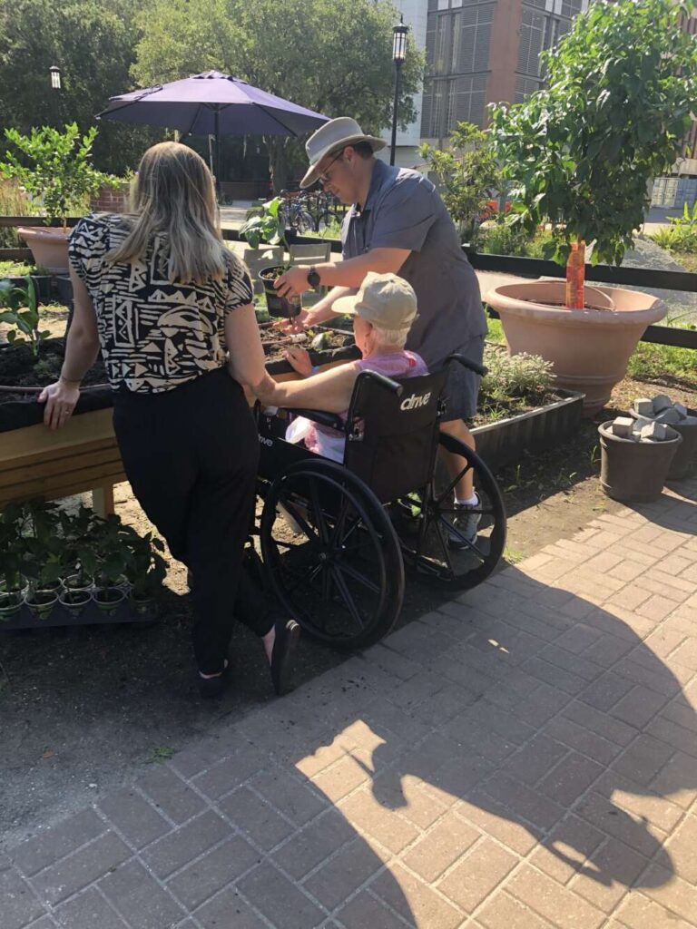 Two people stand and one person sits in a wheelchair, face away from the camera, handling plants in a raised wood garden bed.