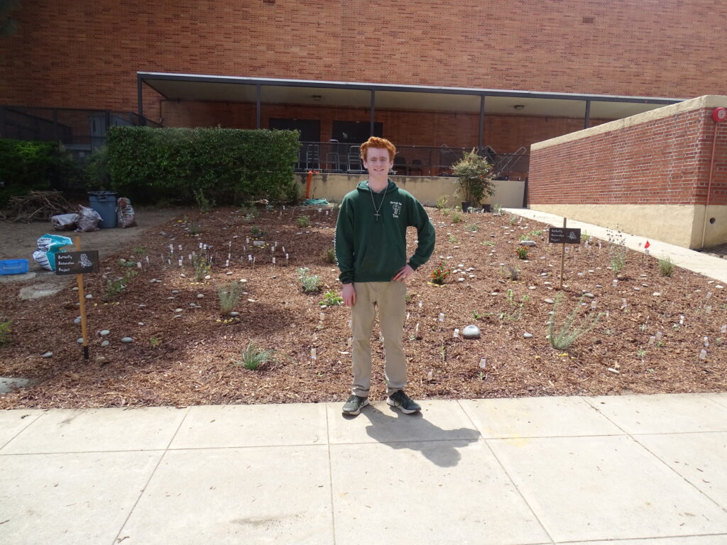 A young adult in a green hoodie and tan pants stands on a sidewalk smiling at the camera, with. In the background are plants on brown barked or mulched ground. Behind that is a red brick building.