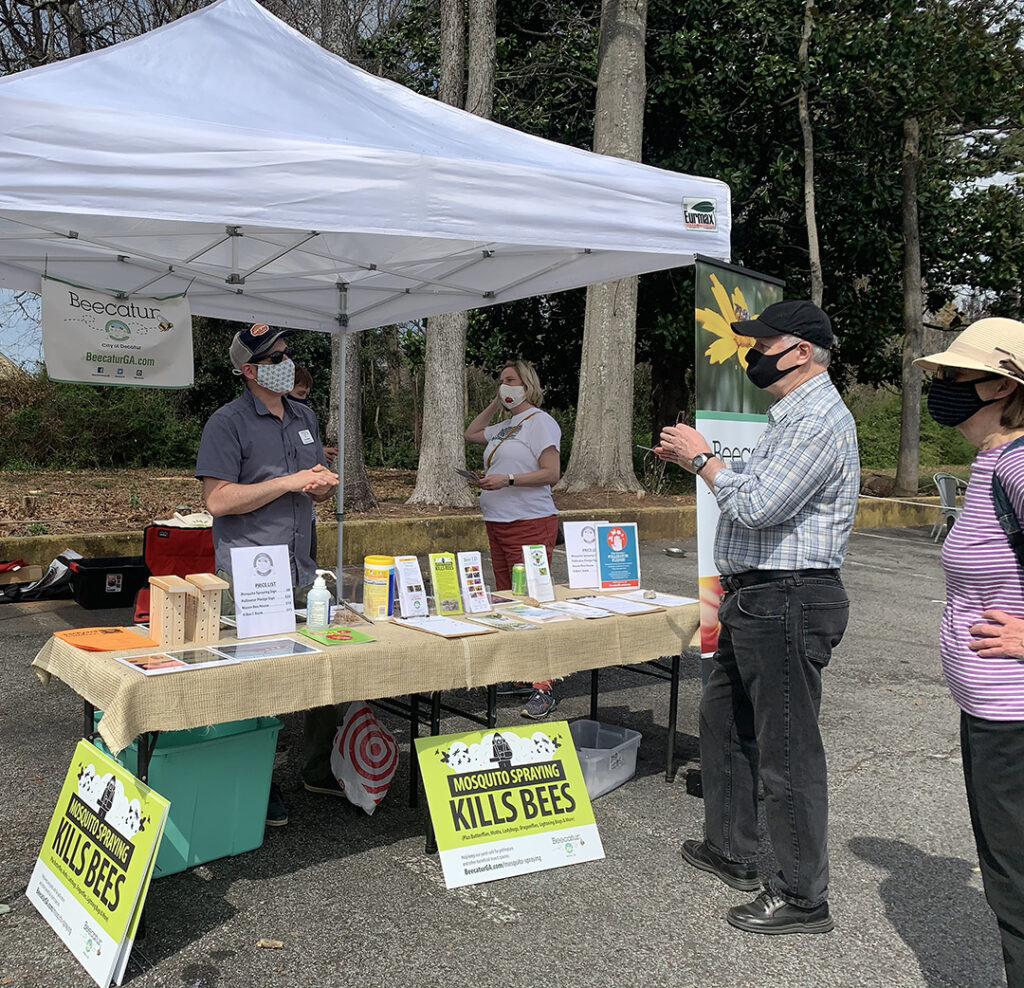 Four people stand by a table under a canopy. Two green and white signs with lack lettering are propped up that say in black text: "Mosquito spraying kills bees".