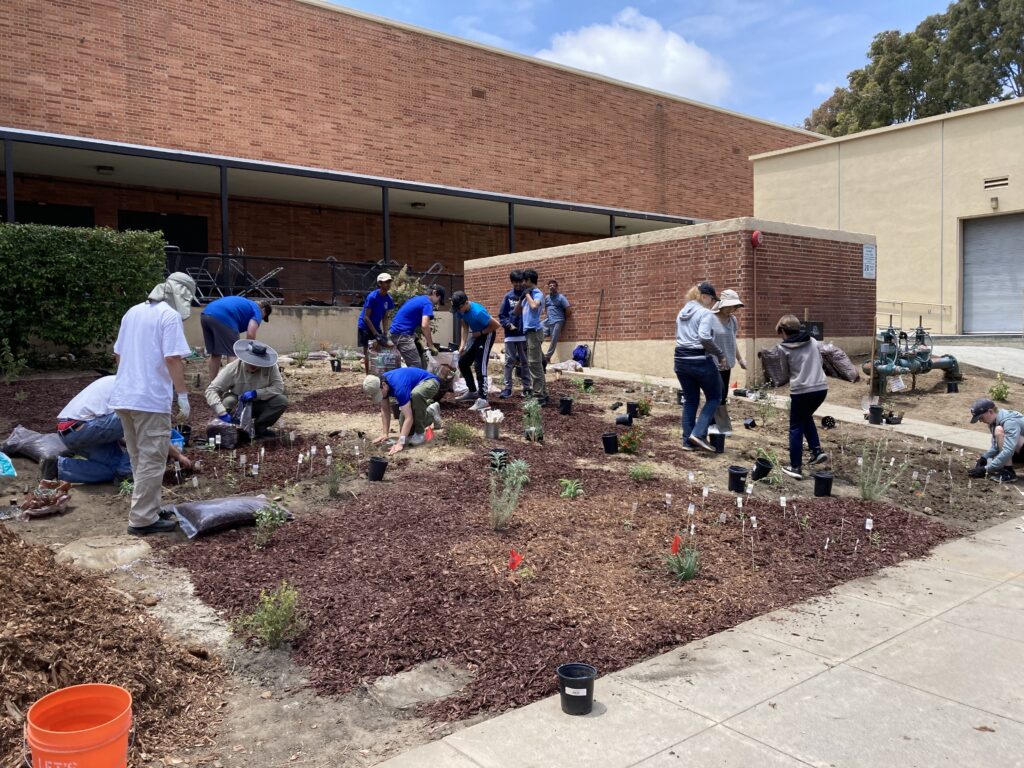 Kids and adults, wearing blue, white, and gray mostly, stand and crouch planting plants in front of a red brick building.