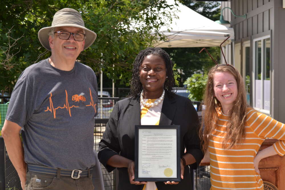 Three people smile facing the camera outside, the middle person holds a framed document.
