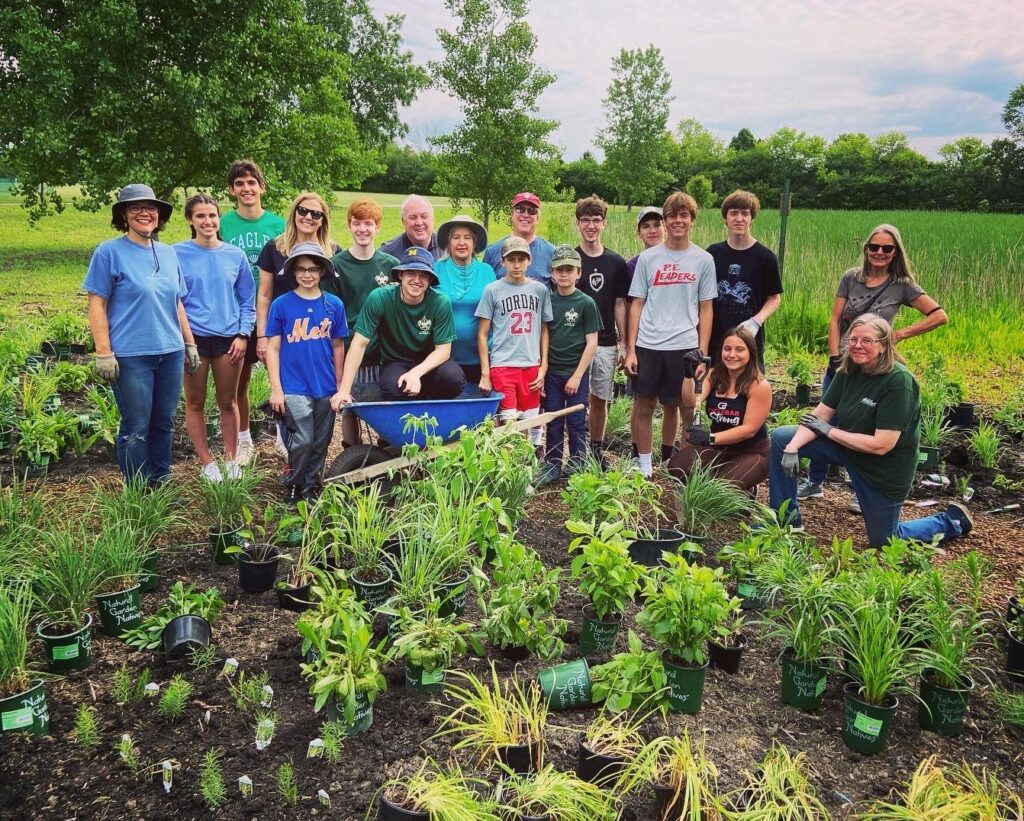 A group of around 20 smiling adults and kids in t-shirts stand in front of a variety of potted and planted plants.