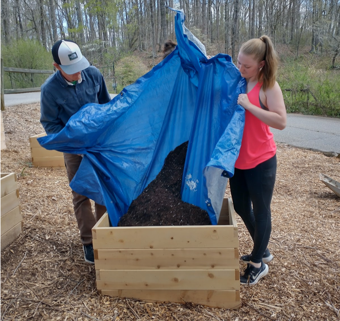 Two students dump soil into a wood raised garden bed, using a large blue tarp.
