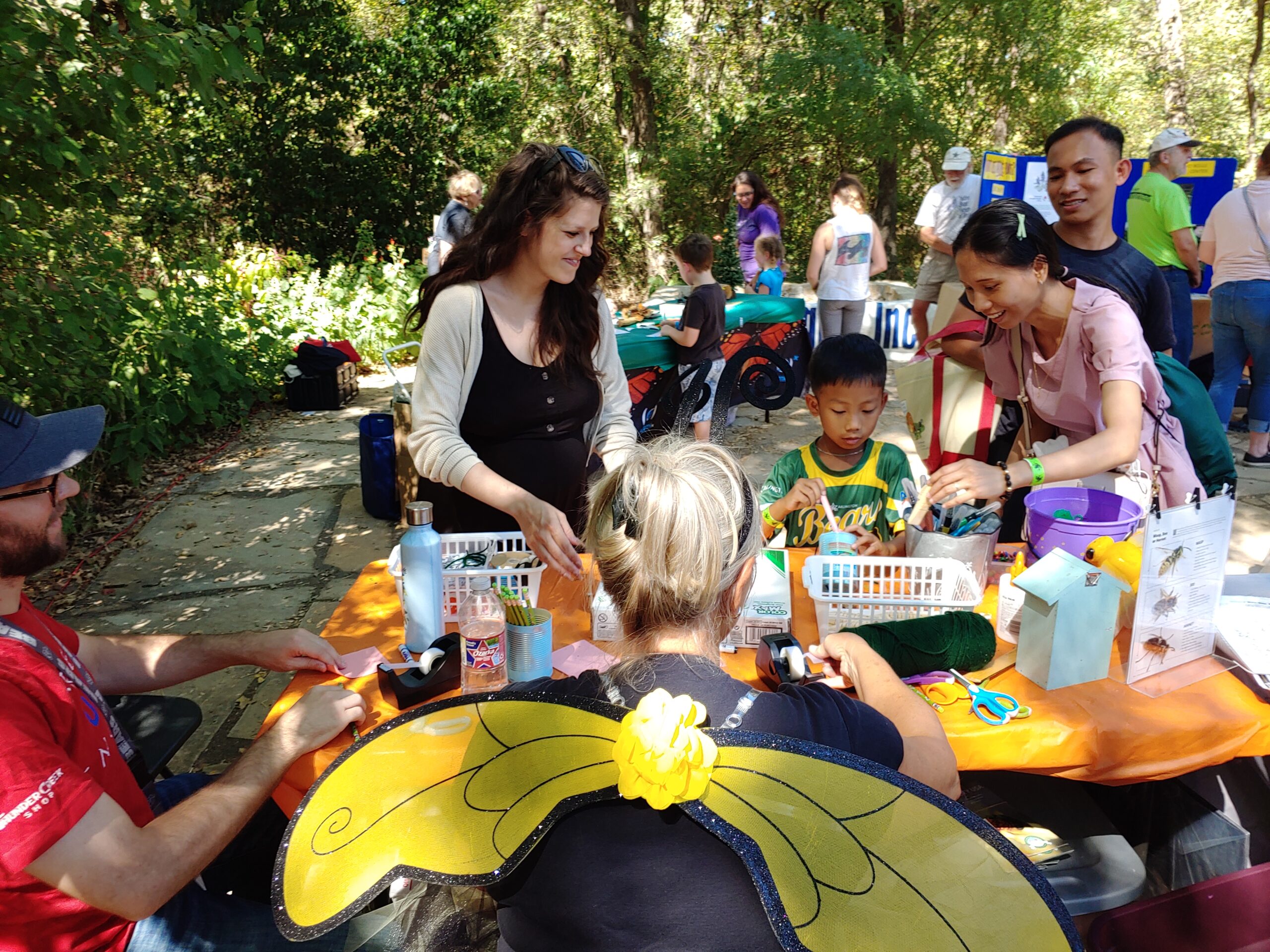 Smiling adults and kids work surround colorful tables outdoors. An adult in a foreground wears large yellow and black wings.