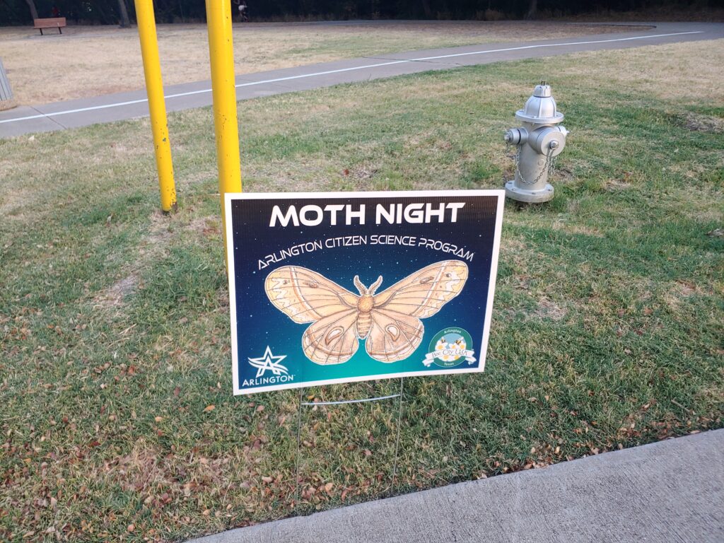 A dark blue lawn sign saying Moth Night with a picture of a moth, on a green lawn
