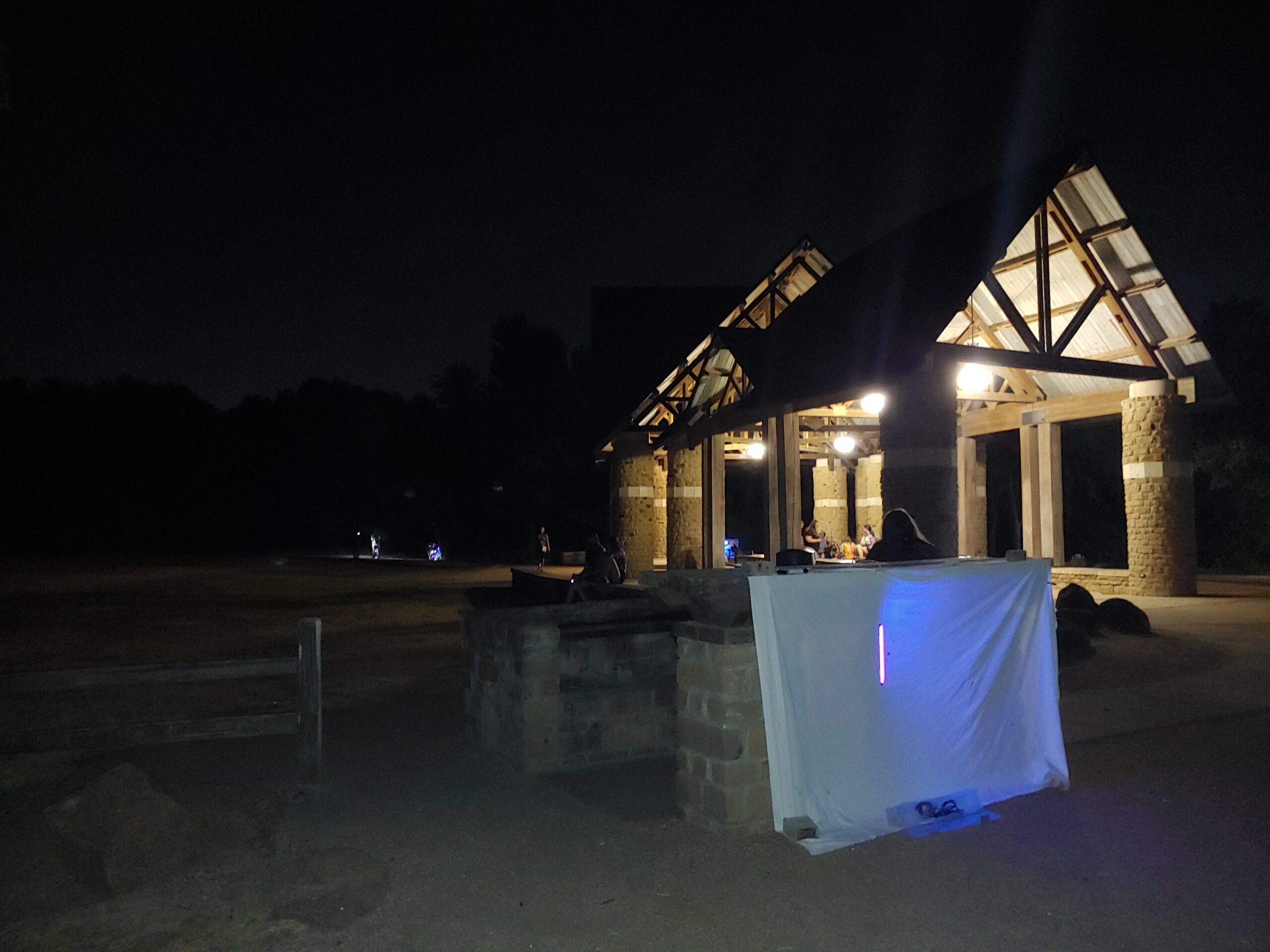 A dark park at night, with a lit shelter in the background. In the foreground, there is a white sheet hung up with a blue night shining on it.