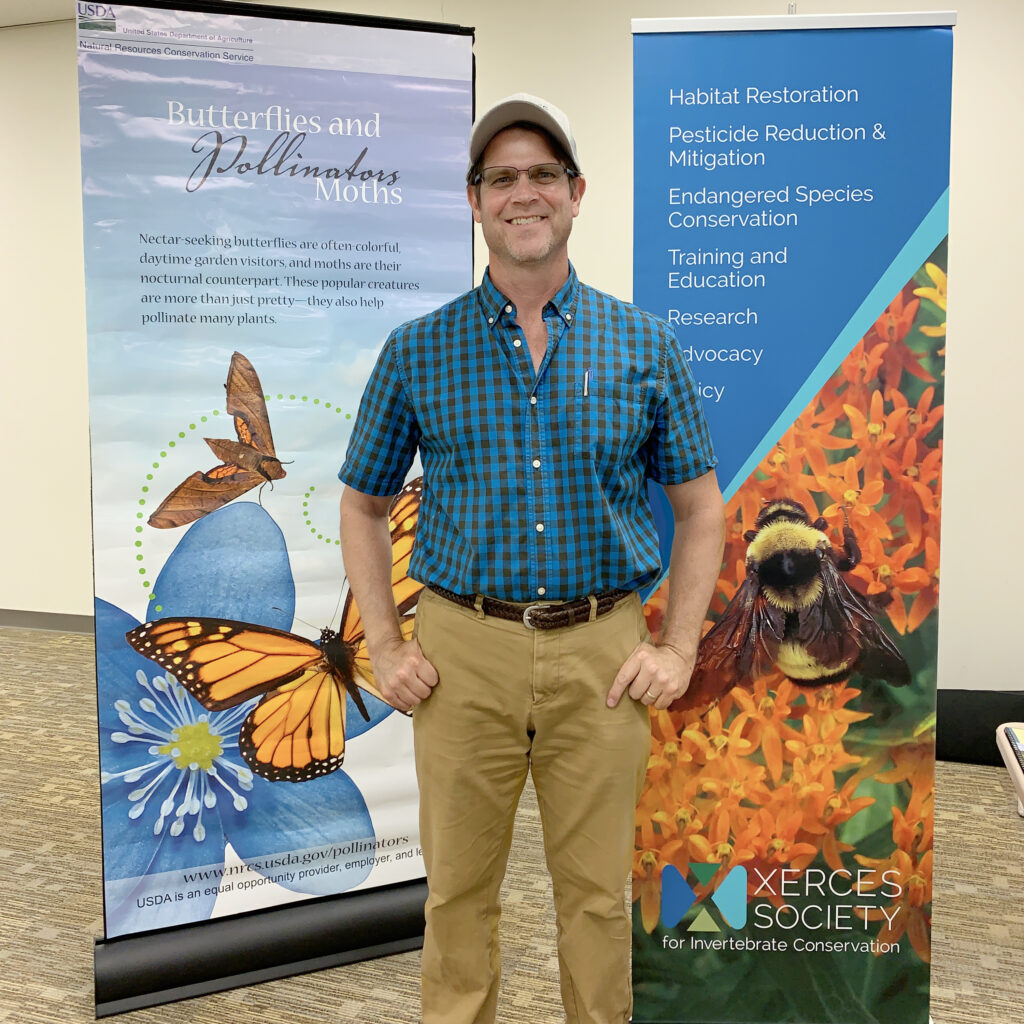 A person stands smiling at the camera in a blue shirt. Light blue and dark blue banners with images of invertebrates are in the background.