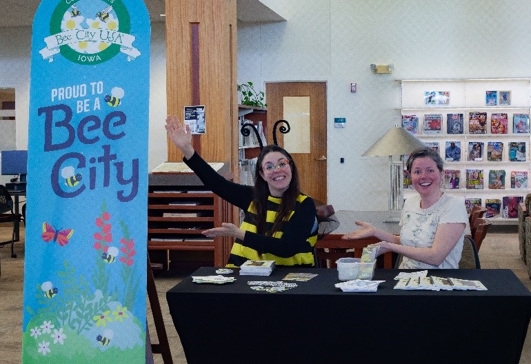 Two people sit behind a table. One of them is in a bee costume and gestures to a large "Proud to be a Bee City" banner.