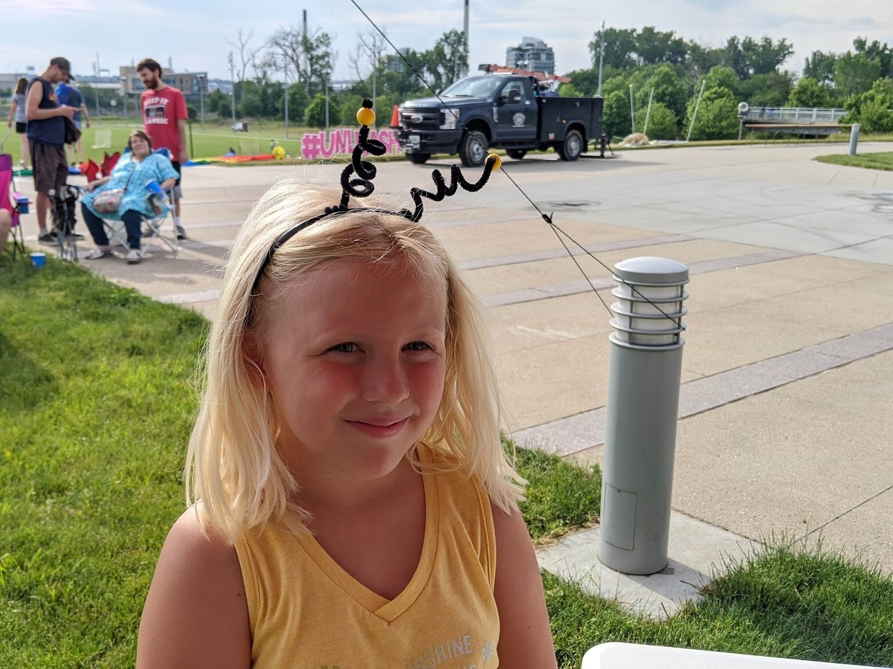 A kid with blonde hair smiles, wearing black spiral pipe cleaner antennae.