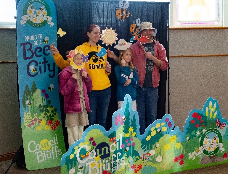 A family poses for a photo holding props (butterflies, watering can, sun, etc), in front of a Bee City-themed backdrop.