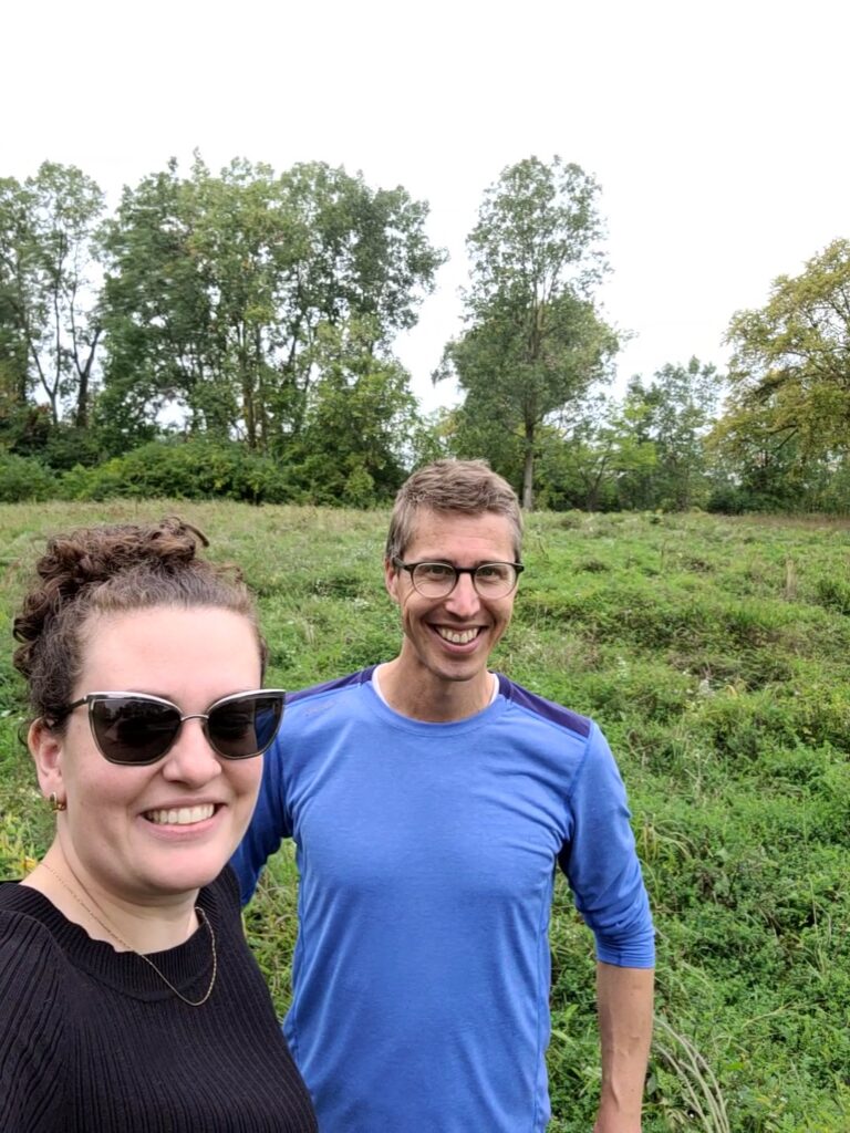 Two people smile at the camera with a green meadow in the background.