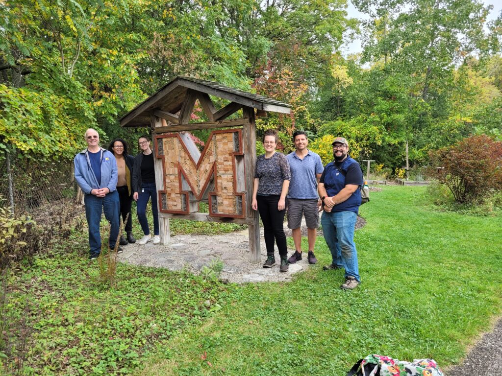 Six people stand in from of a large bee hotel in the shape of an "M".