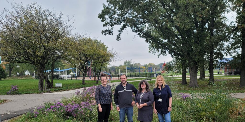 Four adults in darker clothes stand in a row in from of flowers with trees and a playground in the background.