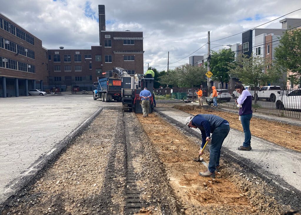 A crew of people remove a strip of asphalt using large equipment, A large brick building is in the background with a gray, mostly cloudy sky above.