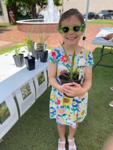 A kid in sunglasses smiles while holding a plant, with a fountain in the background.