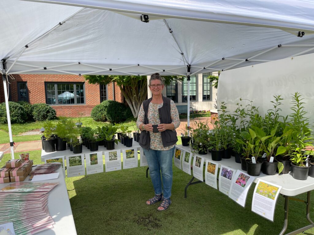 A person in a dark vest and floral shirt stands holding a small plant pot. Standing under a white outdoor canopy, they are surrounded by rows of green plants in black pots on white tables. There is a red brick building in the background.