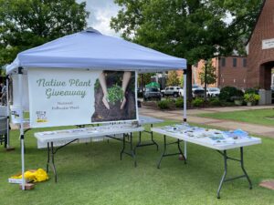 A outdoor canopy with tables set up around it, with a big banner reading "Native Plant Giveaway".