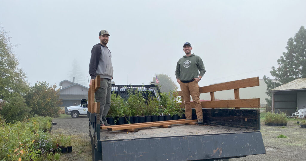 Two smiling people in hoodies stand in the bed of a pickup truck, with a flat of green plants in black pots behind them.