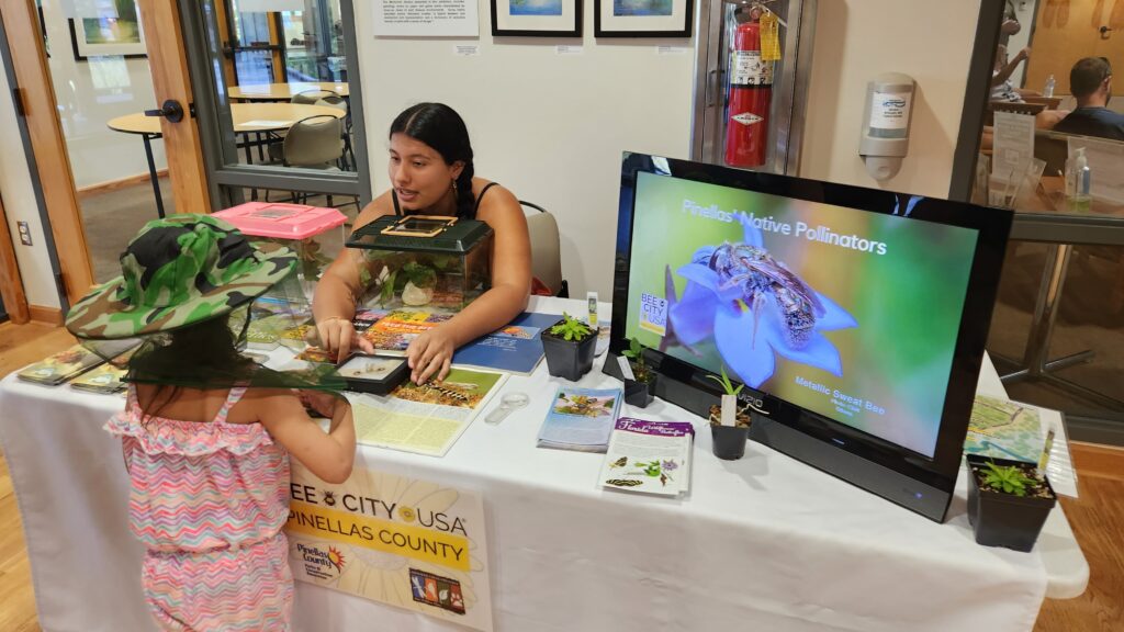 An adult sits behind a display table talking to a child in a beekeeping hat.
