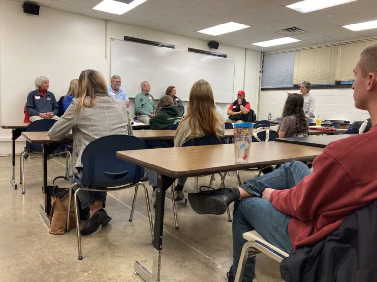 Students in a classroom face people sitting in the front of a classroom, in front of a white board.