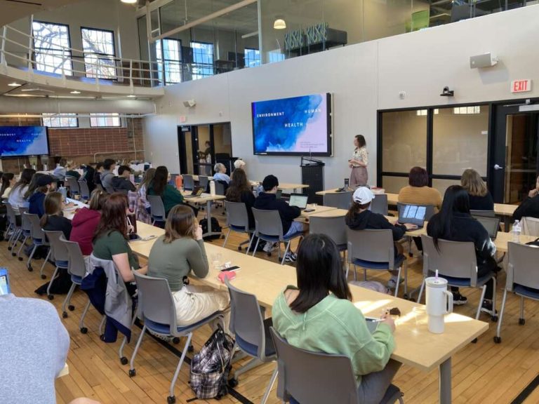 Students at long white tables face a person presenting in a room with a high ceiling.