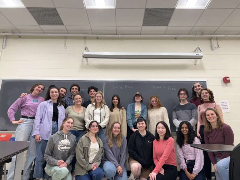 20 smiling people kneel and stand in a group, at the front of a classroom.