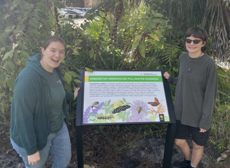 Two people in long sleeves stand smiling on either side of a large interpretive sign, with illustrations and text, with green plants in the behind them.