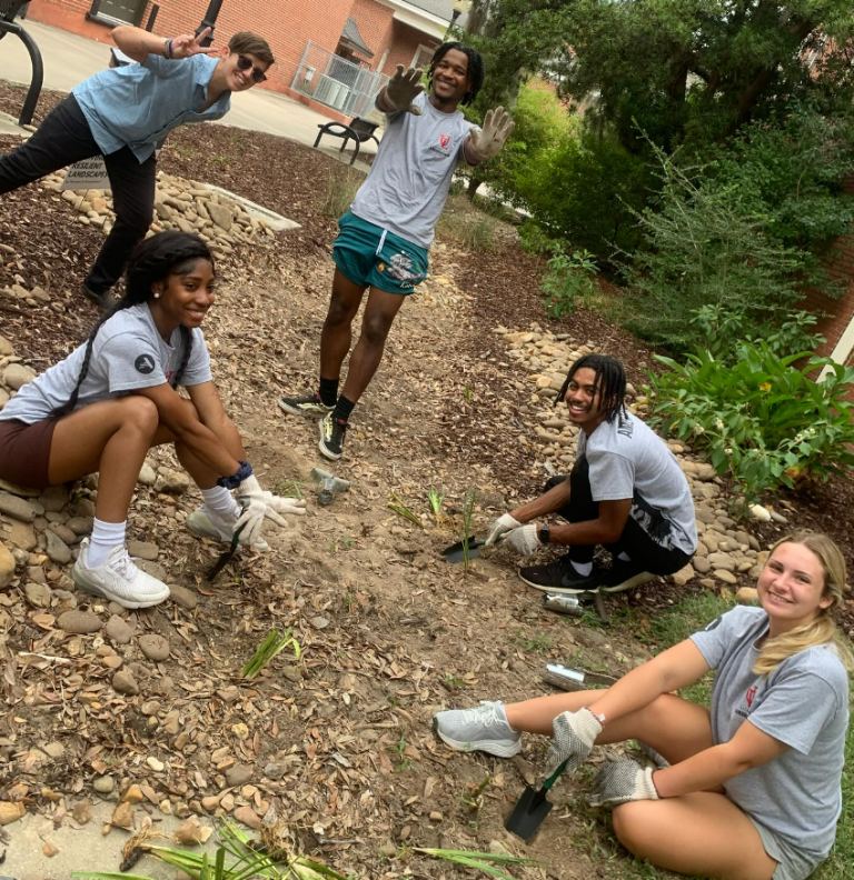 Cheerful people sit and stand, posing for the camera. Some are planting small plants.