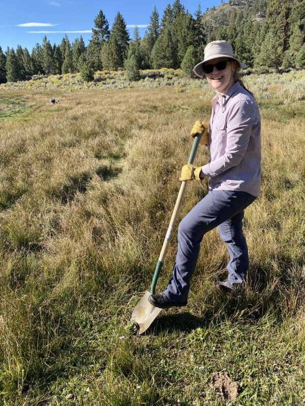 A person with one foot on a shovel smiles at the camera on a sunny day.