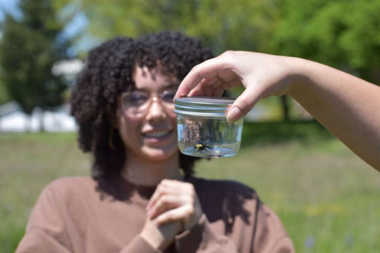 A person smiles looking at a bumble bee in a jar someone is holding up to the camera.
