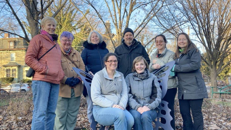 Eight people in warm clothing sit and stand around a butterfly-shaped bench outside with bare trees in the background.