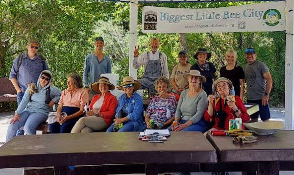 About ten people sit and stand facing the camera smiling, outside, with a banner in the background.
