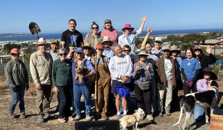 A large group of people pose close together on a sunny day with the ocean in the background.