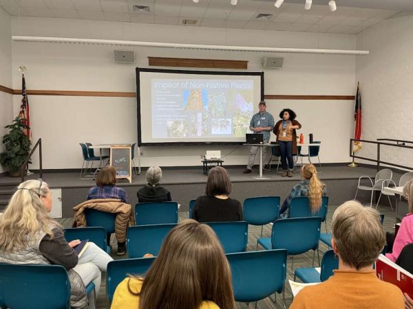 People sit indoors on teal chairs facing towards a person presenting in front of a screen.