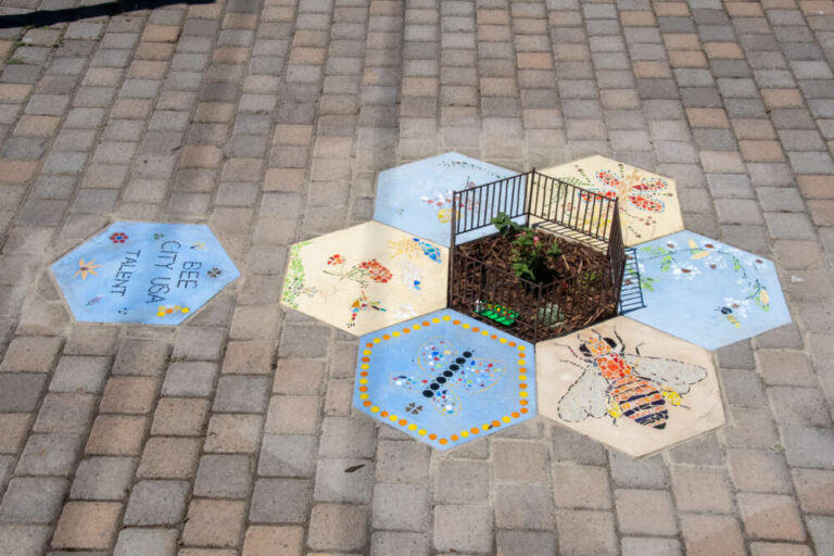 Six hexagon-shaped mosaics on the ground around a hexagon-shaped planter with a tiny fence around it.