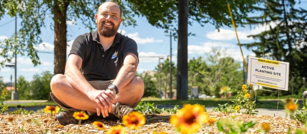 A smiling person in a dark polo shirt sits cross-legged outside in front of red and yellow blanket flowers with a sign reading "Active Planting Site".