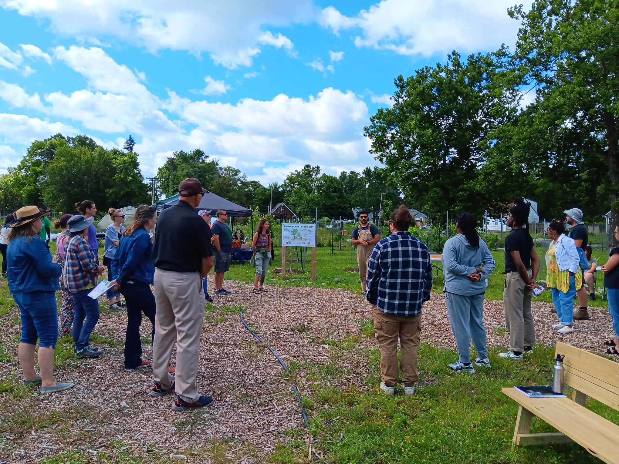 Around 20 people stand in a large circle in front of a fenced garden on a sunny day.