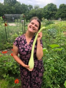 A person in a green garden poses smiling with a long necked light green squash.